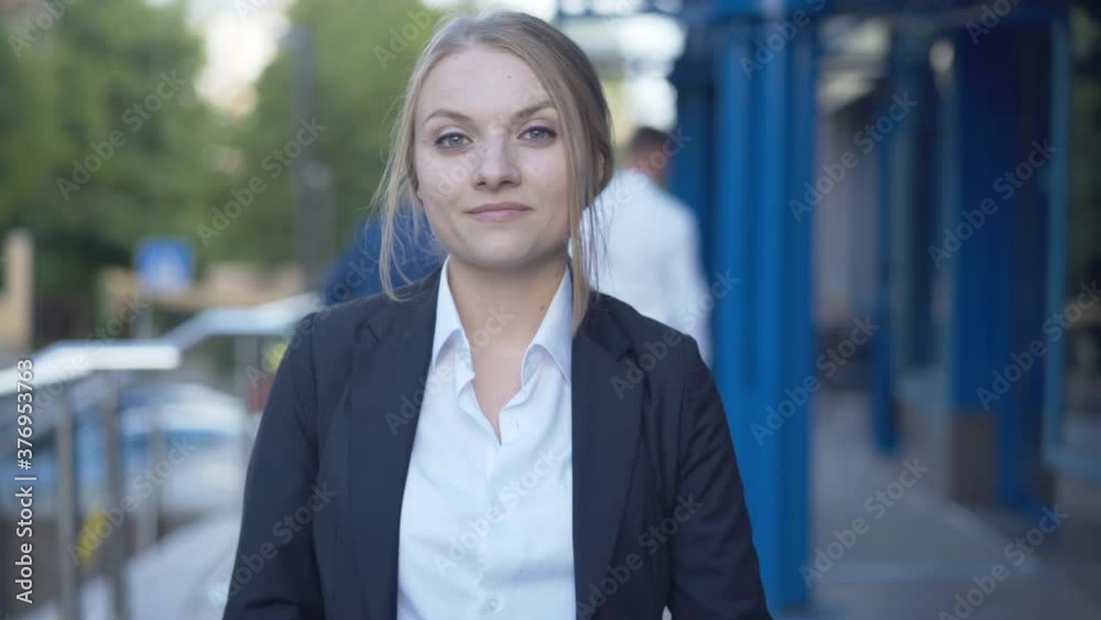 Beautiful young businesswoman posing at business center entrance with blurred people at the background. Portrait of confident smiling Caucasian woman standing outdoors and looking at camera. Beautiful young businesswoman posing at business center entrance with blurred people at the background. Portrait of confident smiling Caucasian woman standing outdoors and looking at camera.