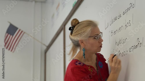 Pretty blond woman teaching spanish in a school on a whiteboard with US flag hanging.