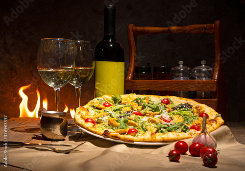 Tasty pizza, bottle of white wine and two wine glasses against the fireplace.