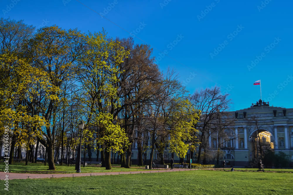 path in the park among the trees with yellow green foliage leaves in the light of the sun on the background of city architecture on the background of blue sky in spring