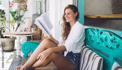 Cheerful young woman reading book and sitting at couch outside