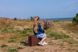 Little blonde girl in denim jacket, blue dress with vintage suitcase and flowers bouquet off-road with sea landscape. Stylish hitchhiker child with long hair on countryside trip. Kid walking outdoors.