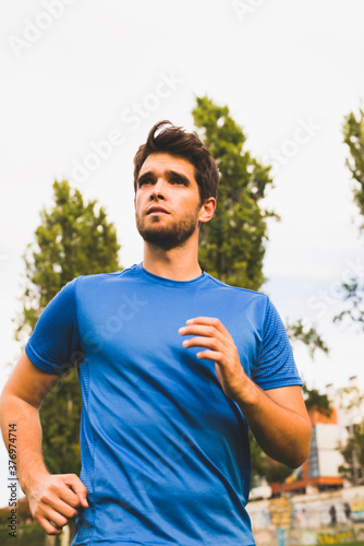 Attractive young man with a beard and blue t-shirt runs through the park in good weather.