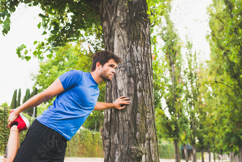 Young man dressed in sportswear performs stretching exercises leaning on a tree in the park. Lifestyle.