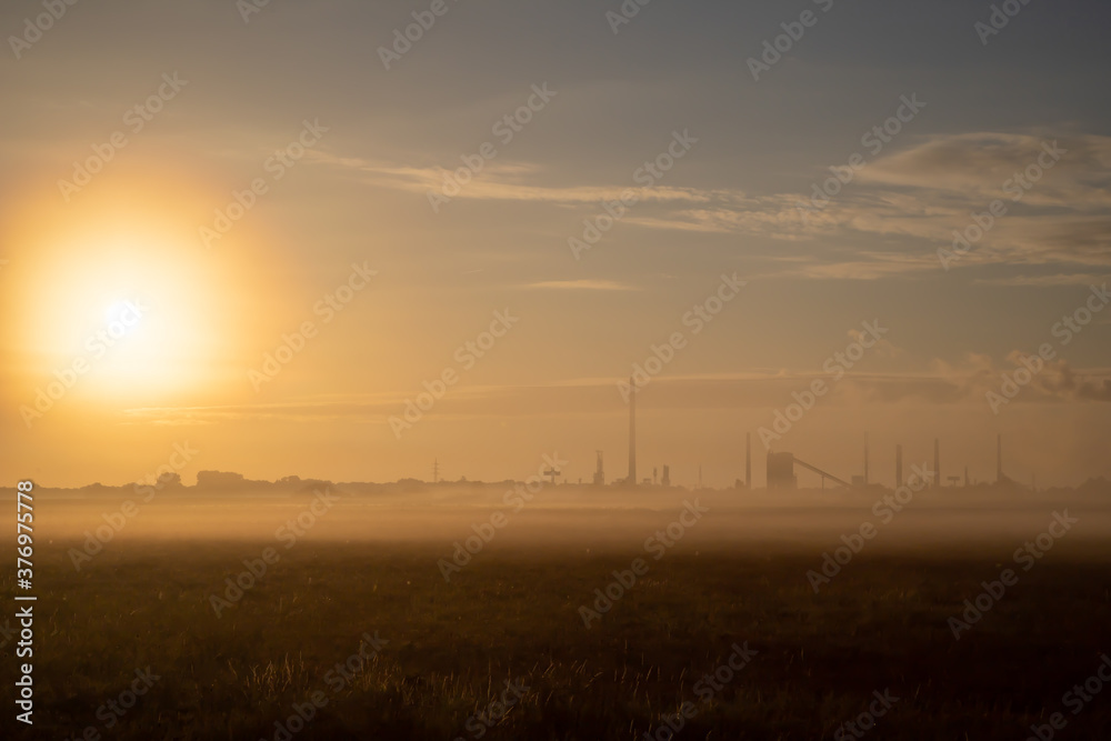 silhouette of a factory with chimneys at dawn, morning fog creates a mystical atmosphere, meadow in the foreground