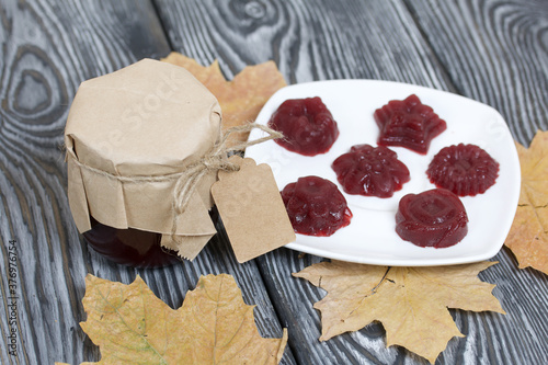 A jar of cherry jam with a label for an inscription and a saucer of cherry marmalade. Dried maple leaves all around. On pine planks painted black and white. photo