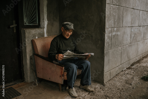 A pensive old man in glasses with gray hair is sitting on an old armchair and reading a newspaper near the house. wrinkles. wisdom. against a dark gray texture wall. camouflage cap. in a knitted sweat