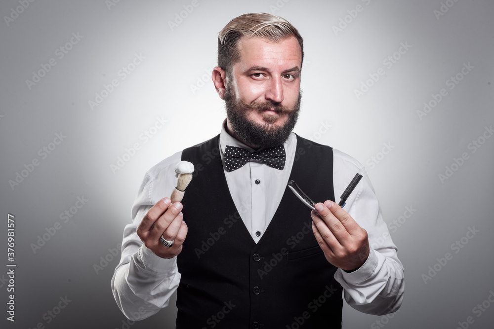 A handsome man in a shirt with a bow tie and a vest holding a blade, a brush with shaving foam. Professional barber concept A handsome man in a shirt with a bow tie and a vest holding a blade, a brush with shaving foam. Professional barber concept