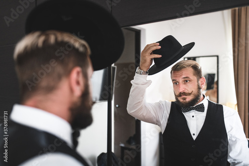 Handsome man with neat beard and mustache takes off his hat in front of the mirror