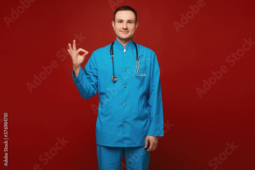 A young man in medical uniform and with a stethoscope showing OK gesture and posing at the red background, isolated. Healthcare and emergency concept photo