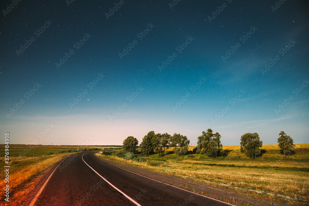 Fototapeta premium Blue Night Starry Sky Above Country Asphalt Road In Countryside And Green Meadow. Night View Of Natural Glowing Stars