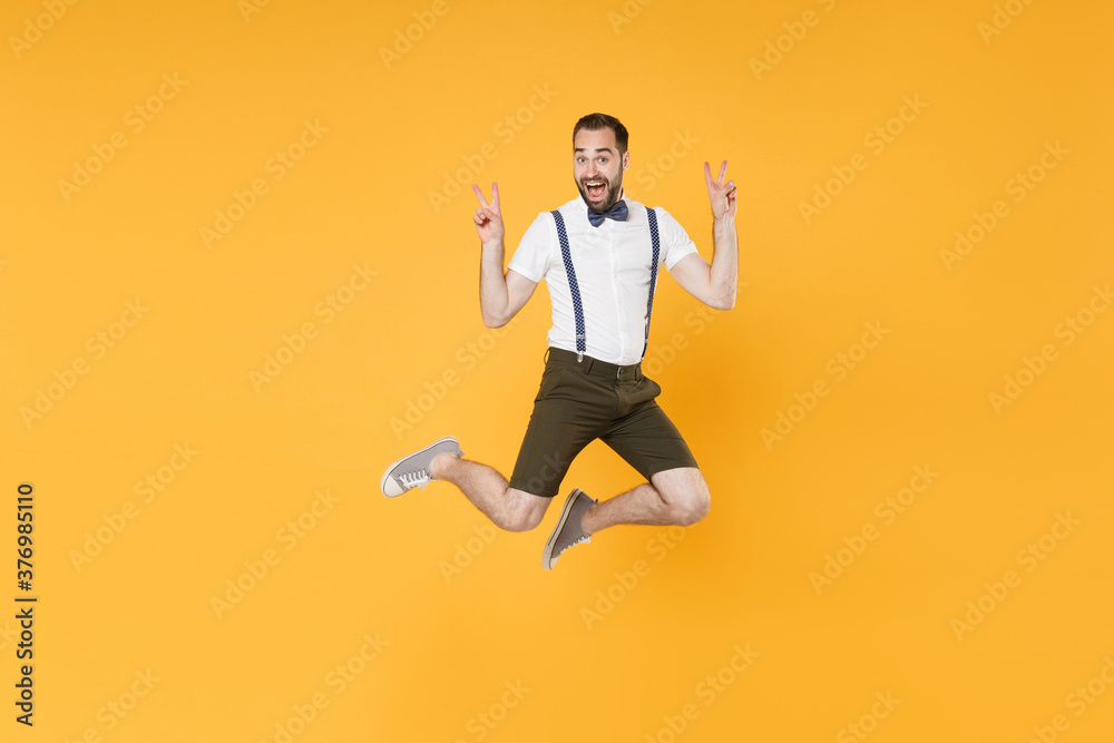 Full length portrait of excited young bearded man 20s wearing white shirt suspender shorts posing jumping showing victory sign looking camera isolated on bright yellow color wall background studio.