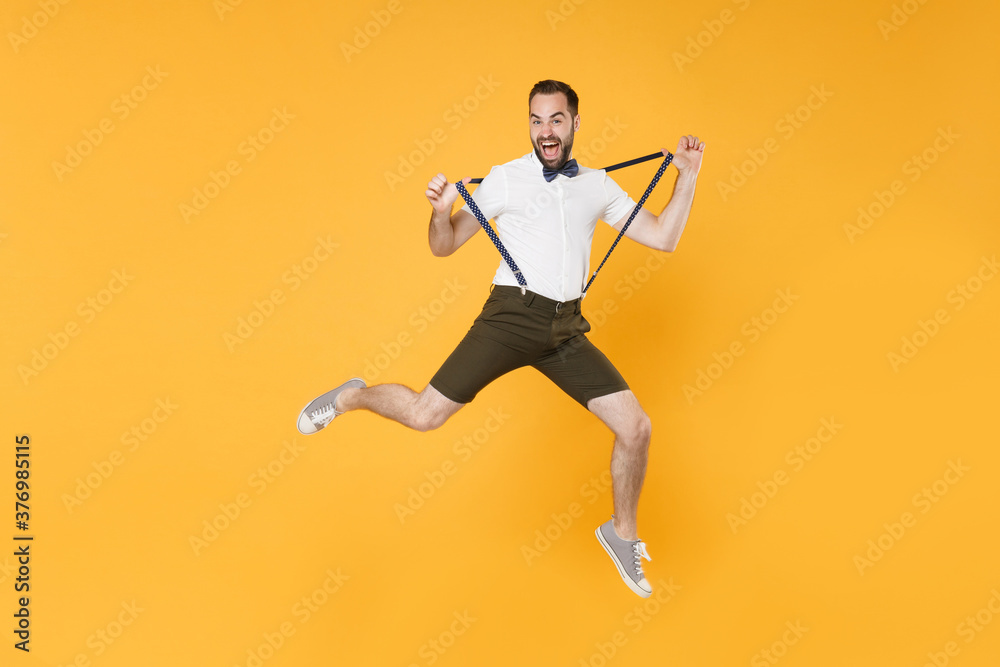 Full length portrait of crazy screaming young bearded man 20s wearing white shirt shorts posing jumping stretching suspender looking camera isolated on bright yellow color wall background studio.