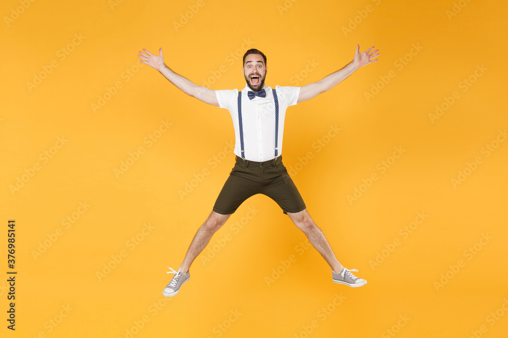 Full length portrait of excited cheerful young bearded man 20s wearing white shirt suspender shorts posing jumping spreading hands and legs looking camera isolated on bright yellow background studio.