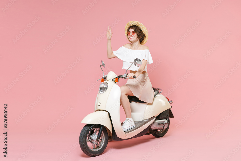 Full length portrait of smiling young woman in white summer clothes hat glasses waving and greeting with hand as notices someone sitting driving moped isolated on pastel pink colour background studio. Full length portrait of smiling young woman in white summer clothes hat glasses waving and greeting with hand as notices someone sitting driving moped isolated on pastel pink colour background studio.