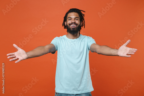 Cheerful young african american man guy in blue casual t-shirt posing isolated on bright orange wall background studio portrait. People sincere emotions lifestyle concept. Reach out stretch hands.