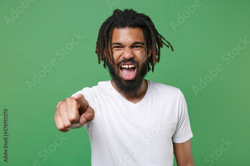 Angry young african american man guy with dreadlocks 20s wearing white casual t-shirt posing pointing index finger on camera screaming swearing isolated on green color wall background studio portrait.