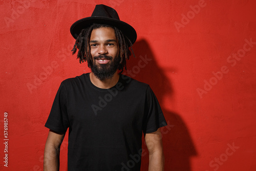 Smiling handsome attractive young african american man guy with dreadlocks 20s wearing black casual t-shirt hat posing looking camera isolated on bright red color wall background studio portrait.