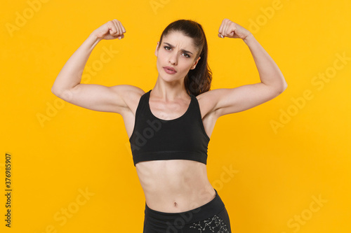 Strong beautiful young fitness sporty woman 20s wearing black sportswear posing working out training showing biceps, muscles looking camera isolated on bright yellow color background studio portrait.