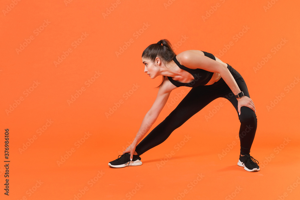 Full length portrait of young fitness sporty woman in black sportswear posing training working out doing stretching exercising for legs looking aside isolated on orange color wall background studio.