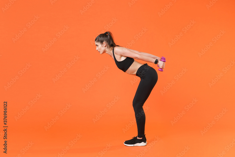 Full length side view portrait of young fitness sporty woman in black sportswear posing training working out doing exercise with dumbbells looking aside isolated on orange color background studio. Full length side view portrait of young fitness sporty woman in black sportswear posing training working out doing exercise with dumbbells looking aside isolated on orange color background studio.