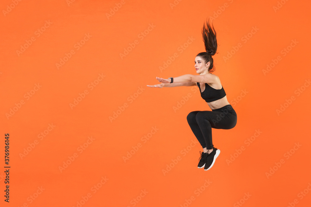 Full length portrait of young fitness sporty woman 20s wearing black sportswear training jumping doing exercise squatting stretching hands looking aside isolated on orange color background studio. Full length portrait of young fitness sporty woman 20s wearing black sportswear training jumping doing exercise squatting stretching hands looking aside isolated on orange color background studio.