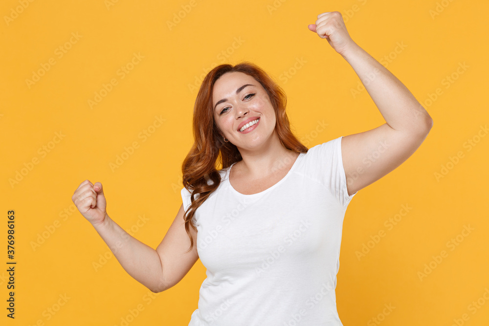 Happy young redhead plus size body positive female woman girl 20s in white blank design casual t-shirt posing clenching fists doing winner gesture isolated on yellow color background studio portrait. Happy young redhead plus size body positive female woman girl 20s in white blank design casual t-shirt posing clenching fists doing winner gesture isolated on yellow color background studio portrait.