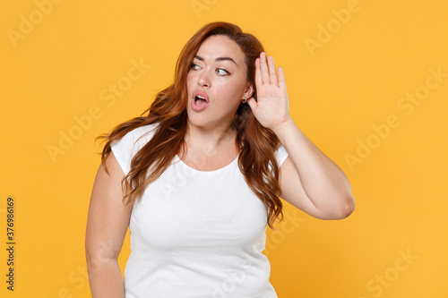 Shocked curious young redhead plus size body positive female woman girl 20s in white casual t-shirt try to hear you overhear listening intently isolated on yellow color background studio portrait.