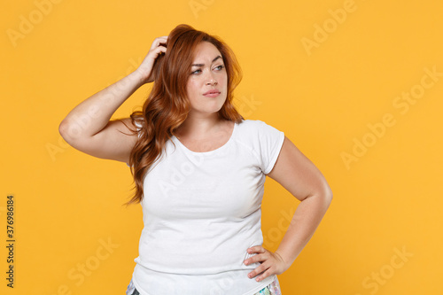 Puzzled pensive young redhead plus size body positive female woman 20s in white blank design casual t-shirt posing put hand on head looking aside isolated on yellow color background studio portrait.