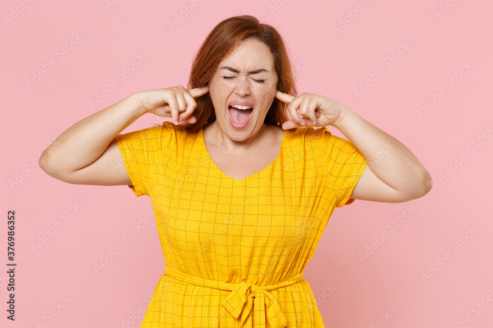 Crazy young redhead plus size body positive female woman girl 20s in yellow dress covering ears with fingers keeping eyes closed screaming isolated on pastel pink color background studio portrait. Crazy young redhead plus size body positive female woman girl 20s in yellow dress covering ears with fingers keeping eyes closed screaming isolated on pastel pink color background studio portrait.