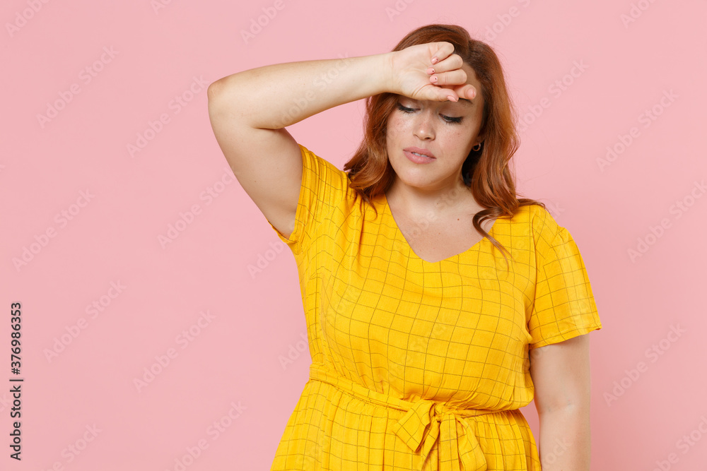 Exhausted tired young redhead plus size body positive female woman girl 20s in yellow dress posing put hand on head looking down isolated on pastel pink color wall background studio portrait.