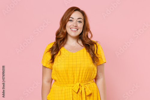 Smiling beautiful attractive young redhead plus size body positive female woman girl 20s in yellow casual dress posing looking camera isolated on pastel pink color wall background studio portrait. photo