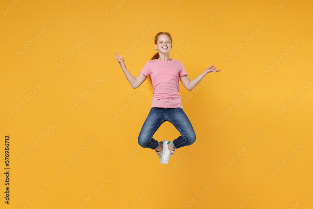 Full length children studio portrait smiling little ginger kid girl 12-13 years old in pink casual t-shirt jumping hold hands in yoga gesture, relaxing meditating isolated on yellow color background.