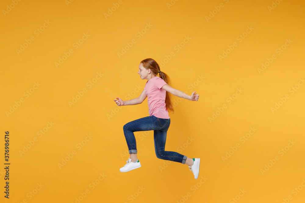 Full length children studio portrait side view of funny little ginger redhead kid girl 12-13 years old wearing pink casual t-shirt posing jumping like running isolated on yellow color wall background.