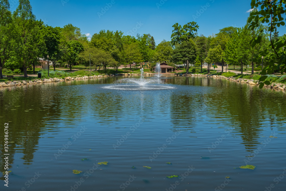 Fountain aerators spray water on blue pond with a stone picnic stand in the background. Sun light reflects off of water droplets suspended mid air in this scenic setting.