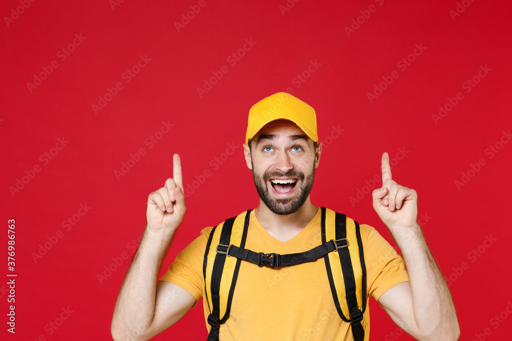 Delivery man 20s in yellow cap t shirt uniform thermal bag backpack points finger up head isolated on red background studio Guy male employee work as courier Service coronavirus covid-19 virus concept