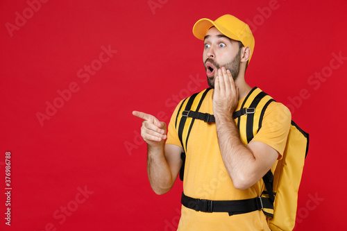 Delivery man 20s in yellow cap t shirt uniform thermal bag backpack points finger aside isolated on red background studio Guy male employee work as courier Service coronavirus covid-19 virus concept
