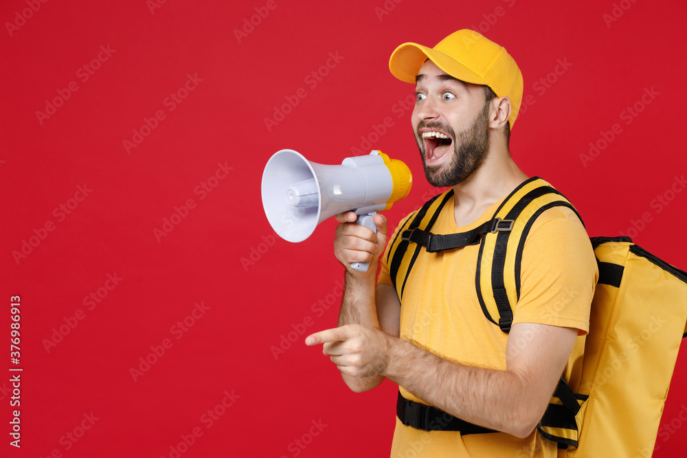Delivery employee man guy male in yellow cap tshirt uniform thermal food bag backpack work courier service during quarantine coronavirus covid-19 scream in megaphone isolated on red background studio Delivery employee man guy male in yellow cap tshirt uniform thermal food bag backpack work courier service during quarantine coronavirus covid-19 scream in megaphone isolated on red background studio