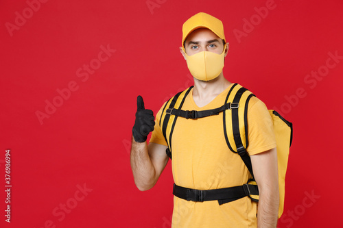 Delivery employee man in yellow cap face mask gloves t-shirt uniform thermal bag backpack with food work courier service during quarantine coronavirus covid-19 virus isolated on red background studio.