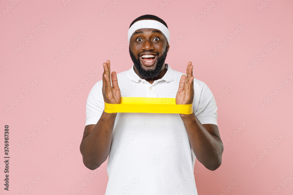 Excited young bearded african american fitness sports man 20s in white headband t-shirt doing exercise with fitness gums spending time in gym isolated on pastel pink color background studio portrait. Excited young bearded african american fitness sports man 20s in white headband t-shirt doing exercise with fitness gums spending time in gym isolated on pastel pink color background studio portrait.