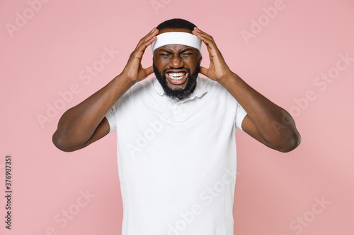Canvas Print Sick displeased young bearded african american fitness sports man in headband t-shirt having headache put hands on head spending time in gym isolated on pink color wall background studio portrait
