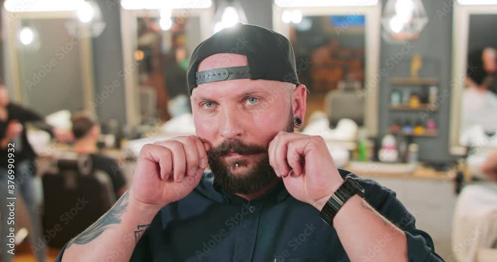 Young bearded and mustashed man with tattoed hands, with chin ring and tunnel earrings in baseball hat, in front of camera curles his mushtashe with one hand in barbershop. In the background the