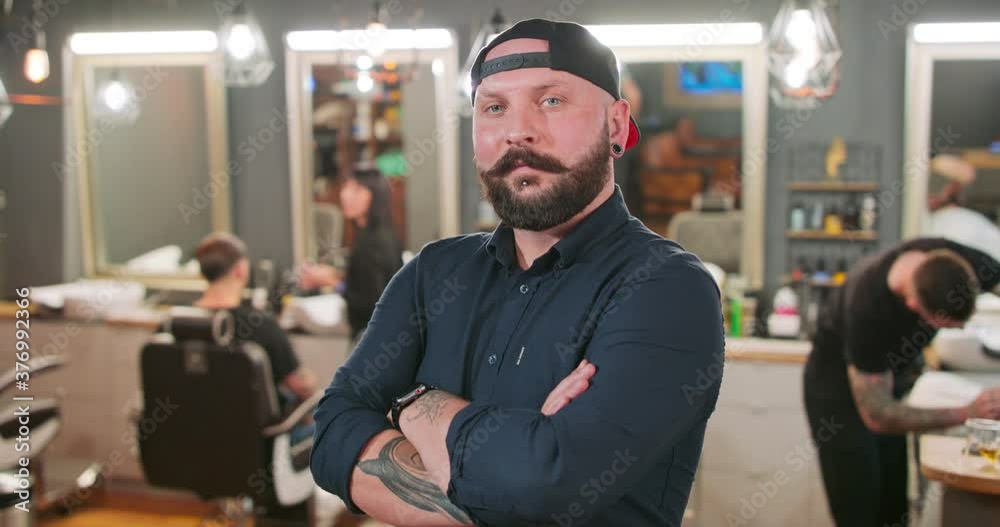 Young bearded and mustashed man, with tattoed hands, with chin ring and tunnel earrings, in baseball hat, in front of camera, crosses his arms over his chest in barbershop. In the background the