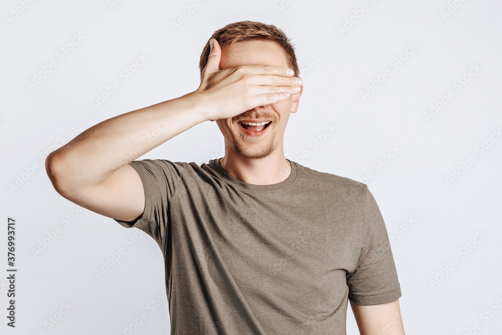 Handsome young man closes his eyes with his hand and smiles. Brunette with a beard in a green shirt posing on a gray background. Place for advertising. I do not see. Handsome young man closes his eyes with his hand and smiles. Brunette with a beard in a green shirt posing on a gray background. Place for advertising. I do not see.