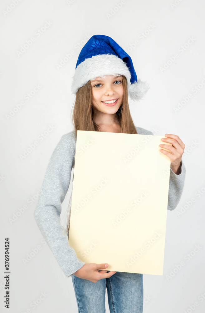 Cute little girl in winter clothes with blue Santa hat posing on light background with blank paper nameplate in hand. Cute little girl in winter clothes with blue Santa hat posing on light background with blank paper nameplate in hand.