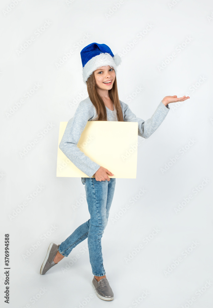 Cute little girl in winter clothes with blue Santa hat posing on light background with blank paper nameplate in hand. Cute little girl in winter clothes with blue Santa hat posing on light background with blank paper nameplate in hand.