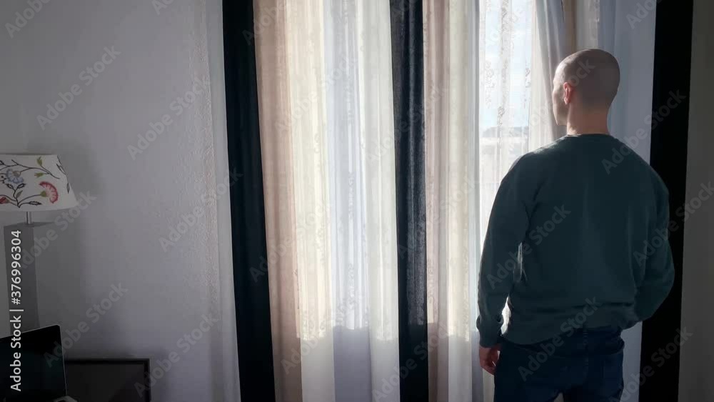 young bald man stands in front of the window, opens the curtains, looks at the street, the concept of business travel, tourism, loneliness, stay at home, quarantine