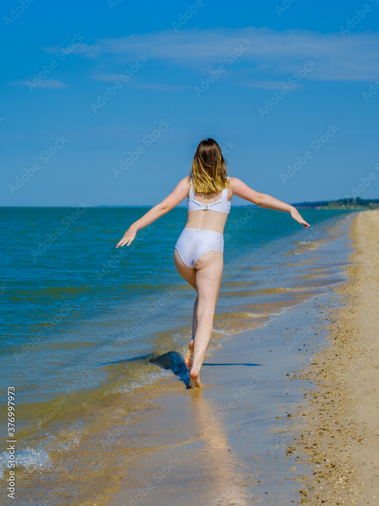 Fototapeta premium Rear view of a girl running away into the distance along a sandy sea beach Splashes of sea water. The girl is wearing a white swimsuit and sunglasses. Freedom. Fitness. Summer sunny day. Copy space