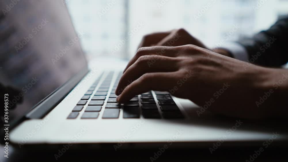 Close-up of hands of unrecognizable businessman typing on laptop keyboard while working at office desk on background of large panoramic window. Concept of office working. Tracking shot in slow motion.