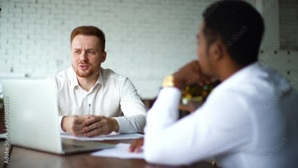 Serious professional African American man with laptop interviewing the caucasian European young candidate for job at successful company. Two business people talking strategy sitting at office desk. Serious professional African American man with laptop interviewing the caucasian European young candidate for job at successful company. Two business people talking strategy sitting at office desk.